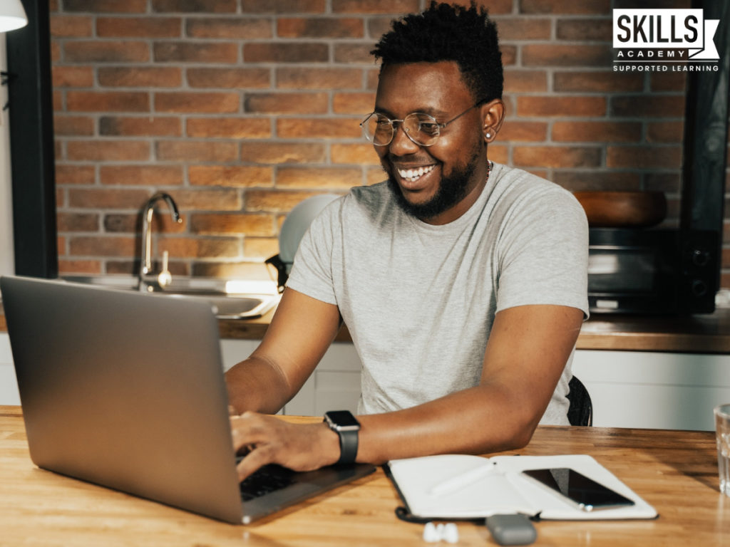 一个穿着灰色毛衣的男人坐在笔记本电脑前微笑。职场技能将帮助你完成工作并保持正轨。