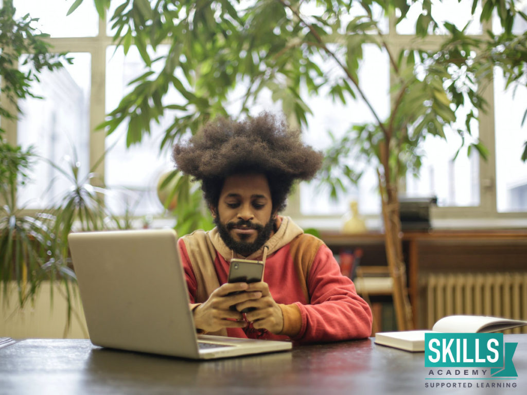 一个年轻人坐在桌前，拿着他的手机和笔记本电脑，看着任何职业的有用技能。