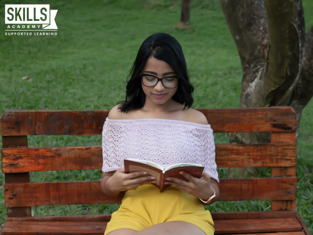 女孩穿着一件白色衬衫和黄色短裤坐在长凳上在公园里阅读。你也可以在任何地方学习当你注册我们的远程教育学院。
