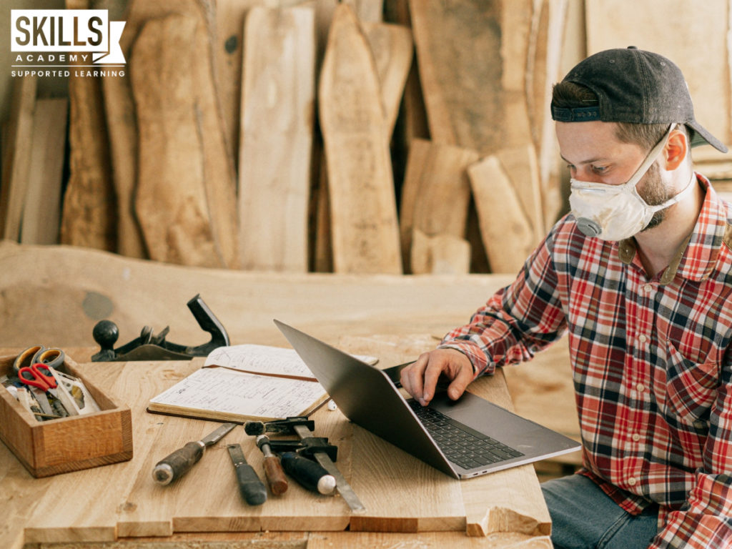 木匠戴着口罩在一台笔记本电脑周围的木头和工具放在桌上。找出什么是你找工作的机会与特定职业课程。