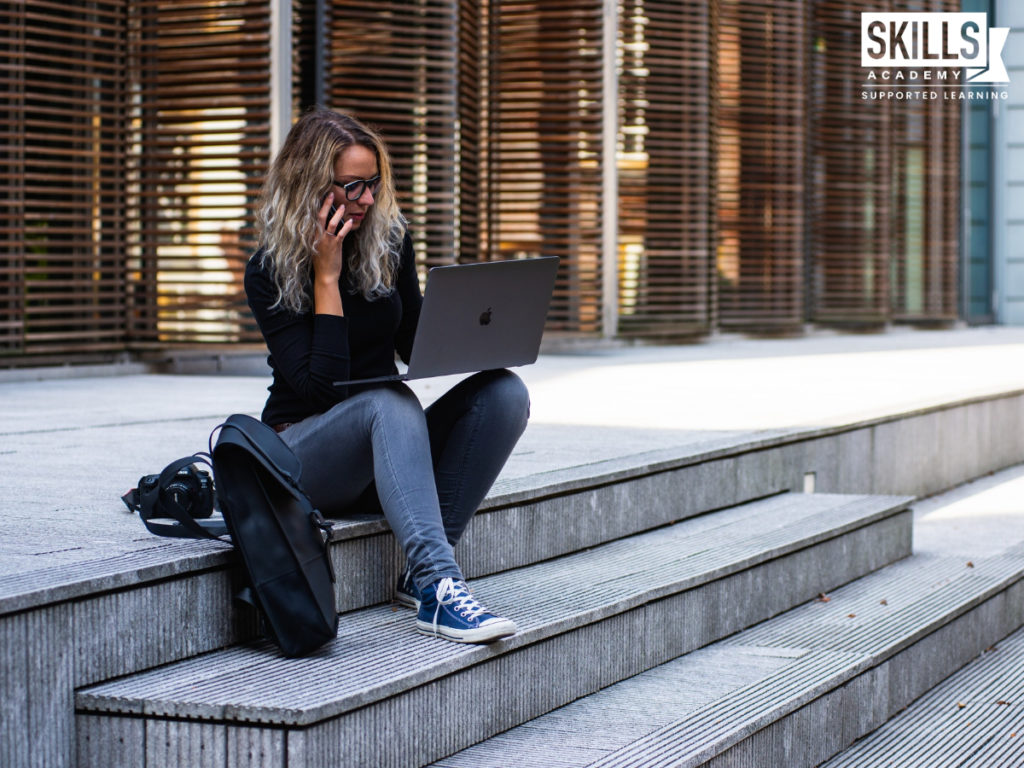一名女子坐在大楼外的台阶上打电话，膝上放着笔记本电脑。开始学习我们的商务英语课程将帮助你为职场做好准备。