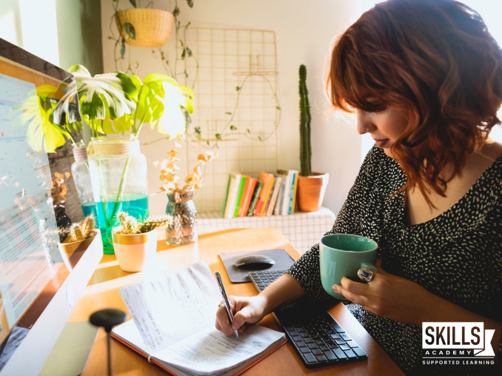 一名女子一边在电脑前的桌子上学习，一边喝着咖啡，一边在书上记笔记。学习如何为远程学习做好准备，创造一个有效的学习空间。